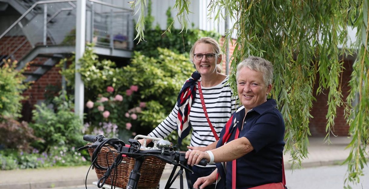 Zwei Frauen mit Fahrrädern lächeln unter einem Baum. Im Hintergrund sind Blumen und ein Gebäude zu sehen.