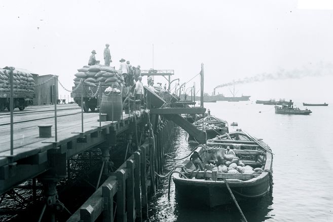 Arbeiter verladen Säcke auf einem Pier, Boote im Wasser, Dampfschiff im Hintergrund.