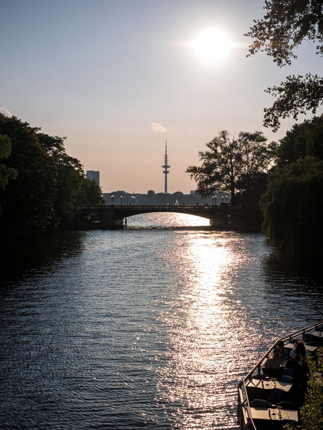 Sonnenuntergang über einem Fluss mit Brücke und Fernsehturm im Hintergrund.