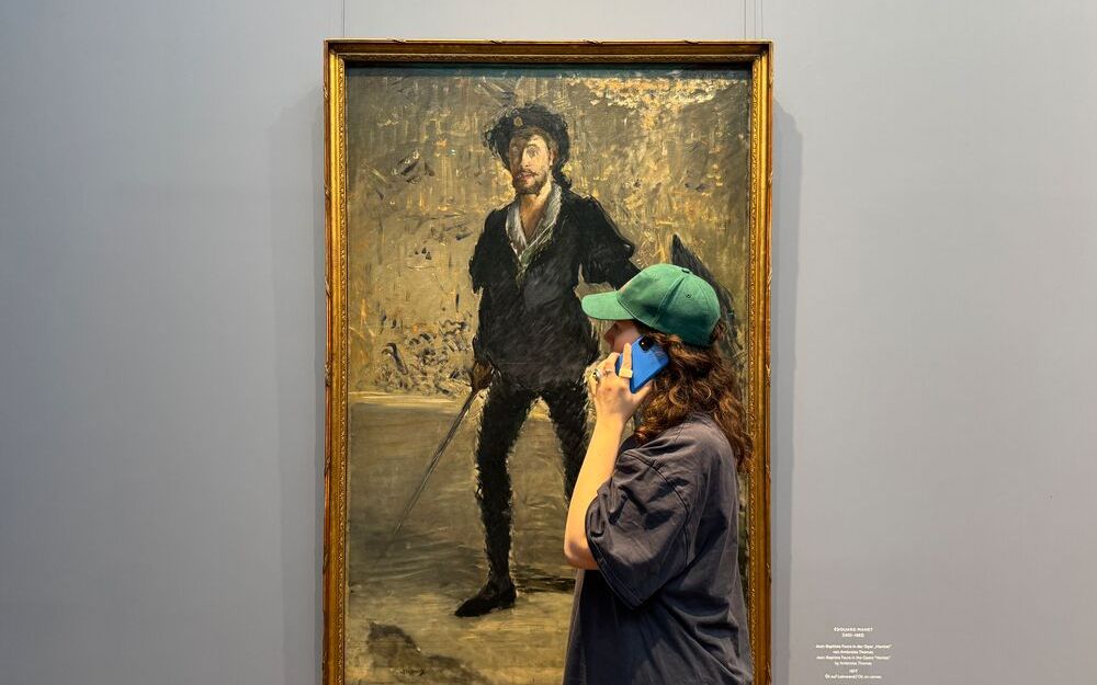 Foto eines Menschen mit langen lockigen Haaren, weitem Hemd und Kappe vor einem Gemälde. Die Person hält ein Handy an das Ohr während sie sich das Gemälde anschaut.