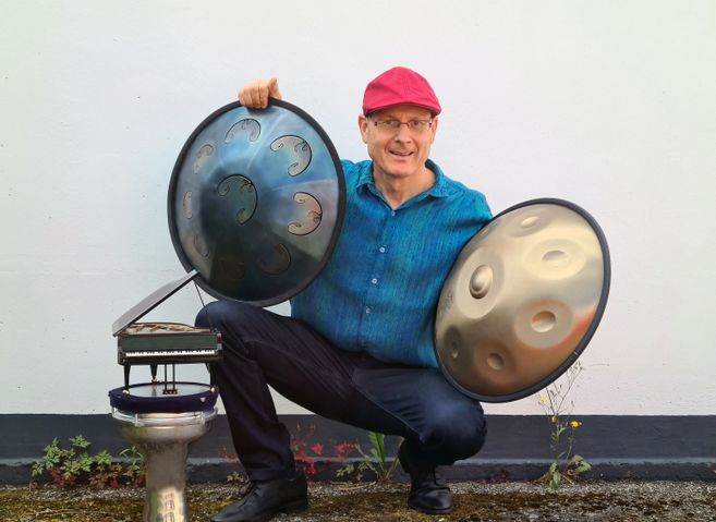 Ein Mann mit roter Mütze hält zwei Handpans und sitzt neben einem kleinen Modell eines Klaviers auf einem Podest.
