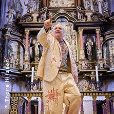 Ein Mann in einem hellen Anzug steht gestikulierend vor einem kunstvoll verzierten Altar in einer Kirche.