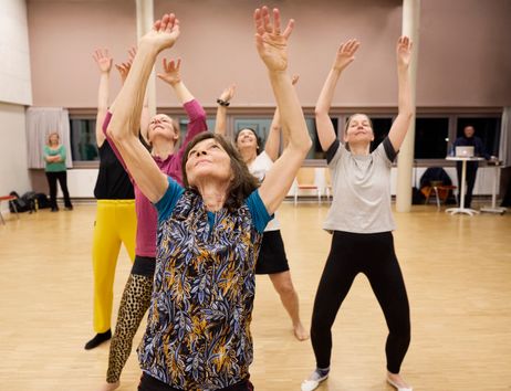 Fünf Personen stehen in einem Tanzstudio hintereinander und haben je beide Arme in Richtung Decke gestreckt. Sie tragen bunte Sportkleidung und ihre Blicke sind in Richtung Decke gerichtet.