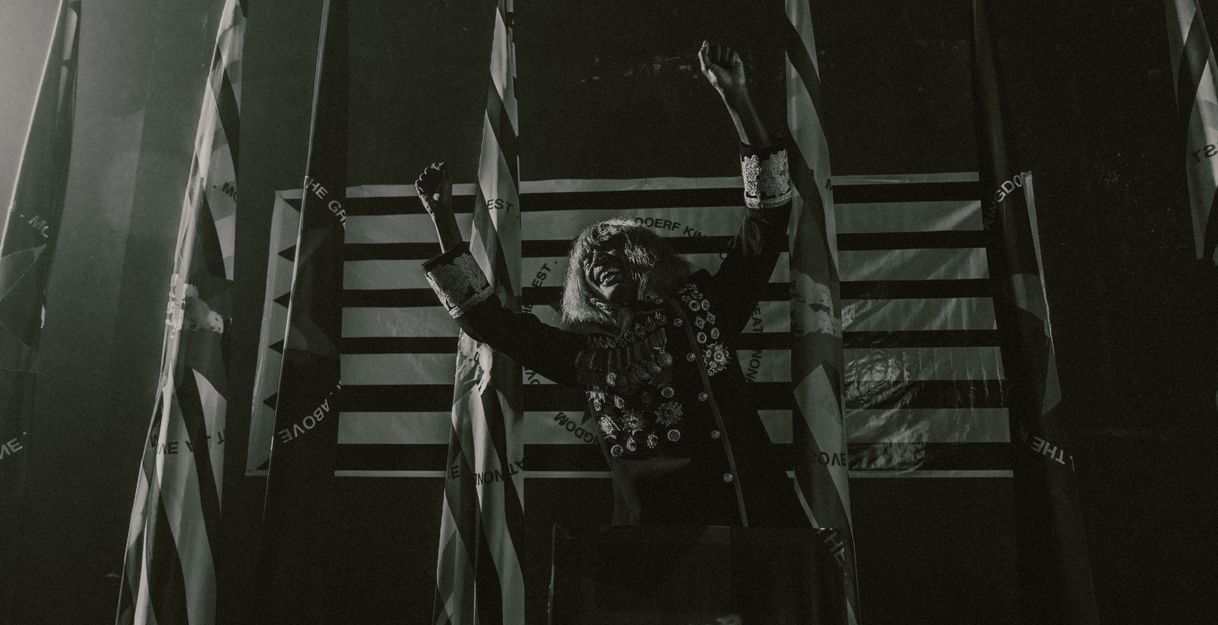 KABEAUSHÉ steht in einer Uniform mit vielen Orden auf einer Bühne vor vier Flaggen, streckt die Arme seitlich nach oben und lacht.