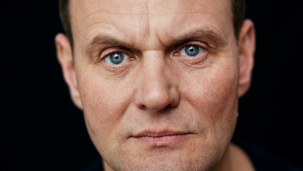 Nahaufnahme eines Mannes mit blauen Augen und ernstem Gesichtsausdruck vor schwarzem Hintergrund.