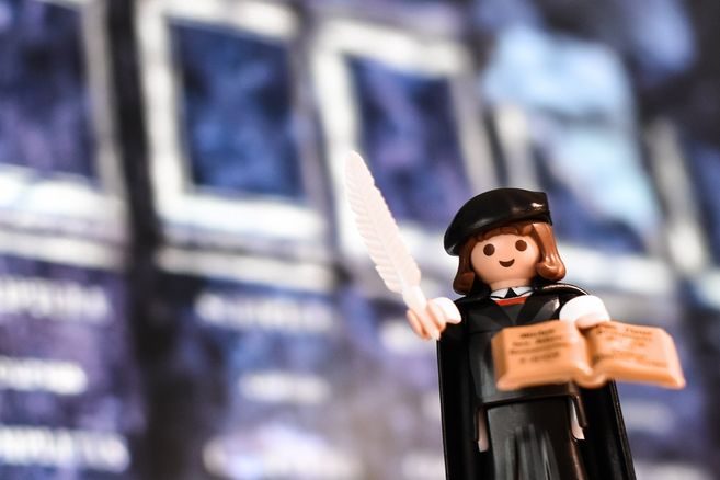 Playmobil-Figur in historischer Kleidung mit Feder und Buch vor unscharfem Hintergrund.