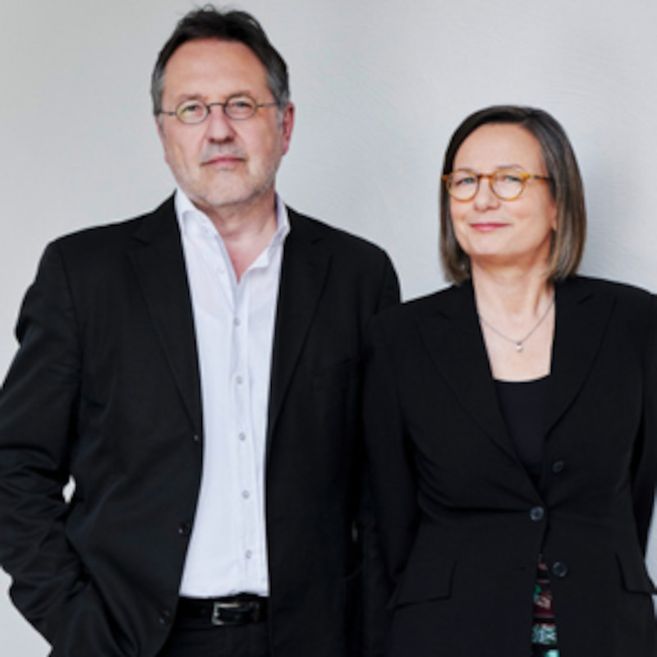 Ein Mann und eine Frau in formeller Kleidung stehen nebeneinander vor einem neutralen Hintergrund.