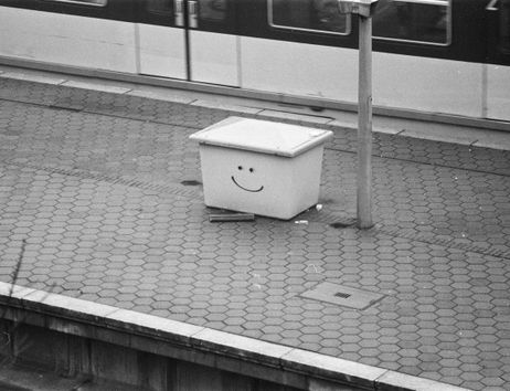 Auf einem Bahnsteig steht ein kasten, auf den ein Smiley gesprüht ist. Im Hintergrund fährt eine alte S-Bahn. Das Bild ist aus den 90ern und in schwarz-weiss.