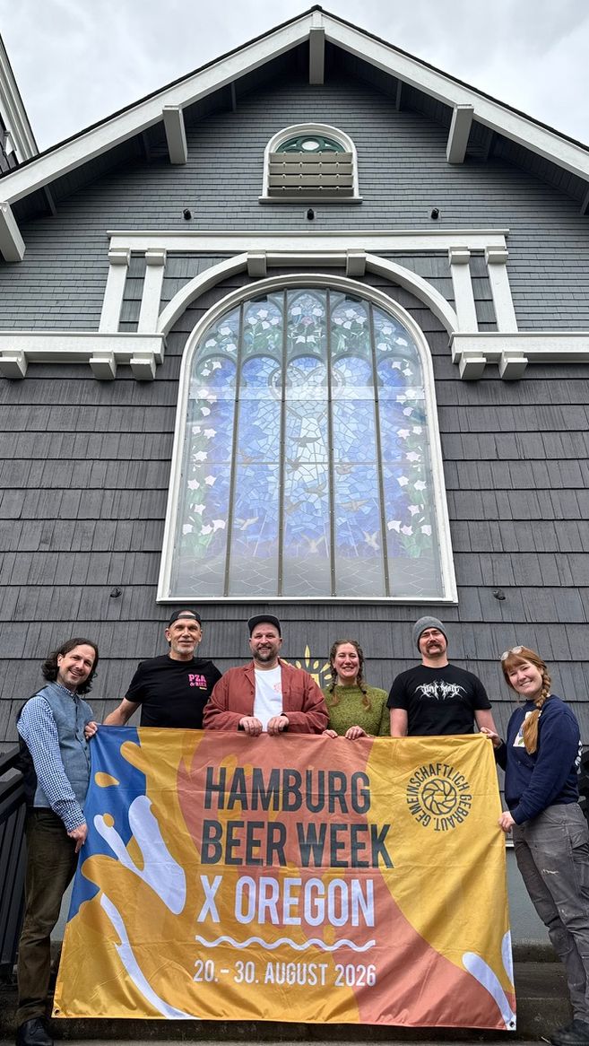 Gruppenfoto der Hamburg Beer Week x Oregon Reise: Sechs Personen stehen vor einer ehemaligen Holzkirche mit großem Fenster und halten ein Banner zur Hamburg Beer Week x Oregon 2026.