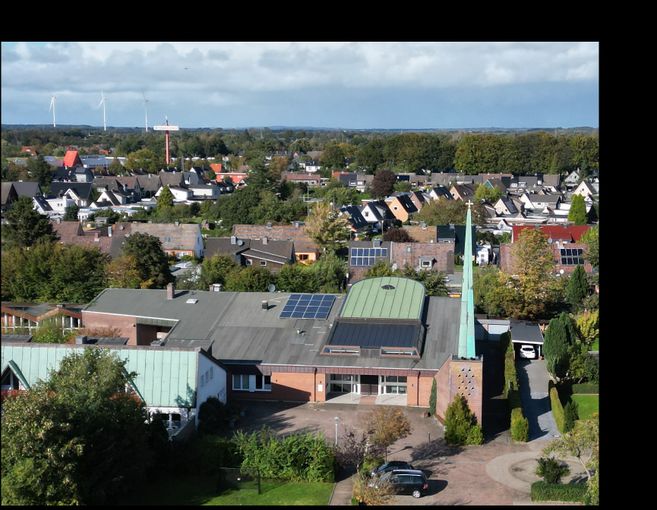 Blick über ein Wohngebiet auf eine Kirche mit grünem Turm und Kreuz; im Hintergrund Windräder und bewölkter Himmel.
