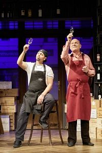 Zwei Männer in Schürzen halten Weingläser hoch, als ob sie den Wein begutachten. Sie stehen in einem Raum mit Weinflaschen im Hintergrund.