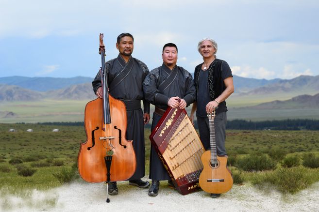 Drei Musiker stehen in traditioneller Kleidung mit Instrumenten vor einer weiten Landschaft.