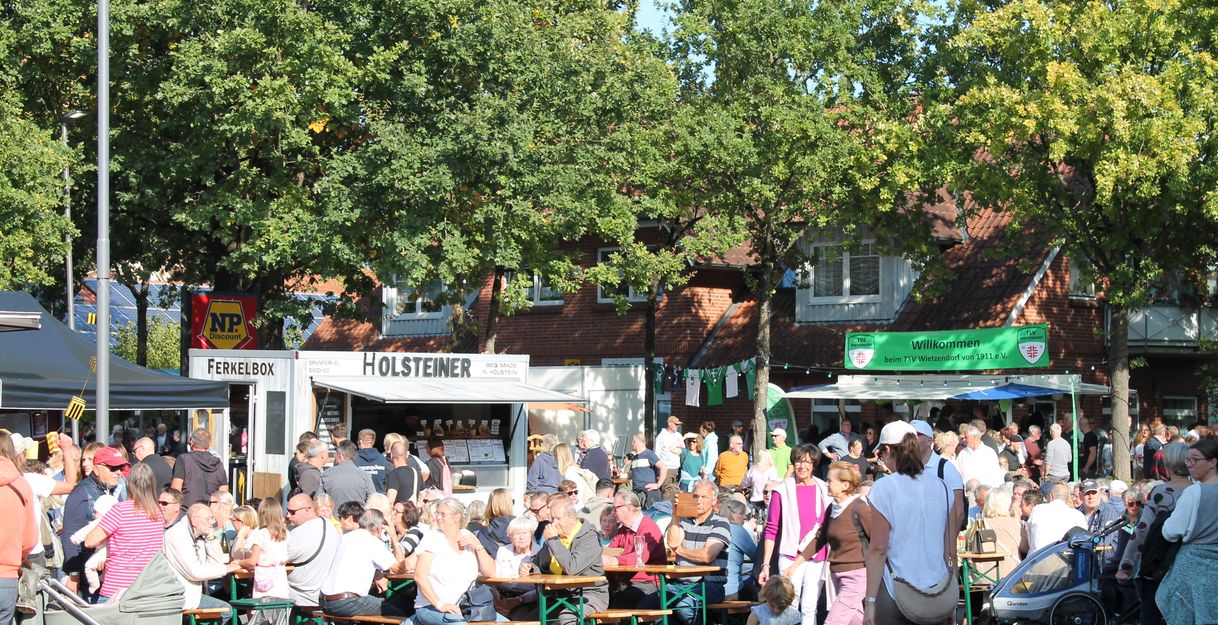 Menschenmenge bei einem Fest in Wietzendorf mit Essensständen und Biertischen unter Bäumen.
