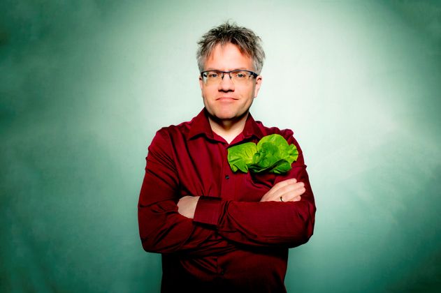 Bild von Matthias Reuter im weinroten Hemd und mit Salatblatt in der Hemdtasche, grüner Hintergrund. 
