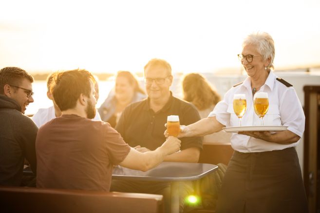 Eine Frau serviert Bier an eine Gruppe von Menschen auf einem Boot bei Sonnenuntergang.