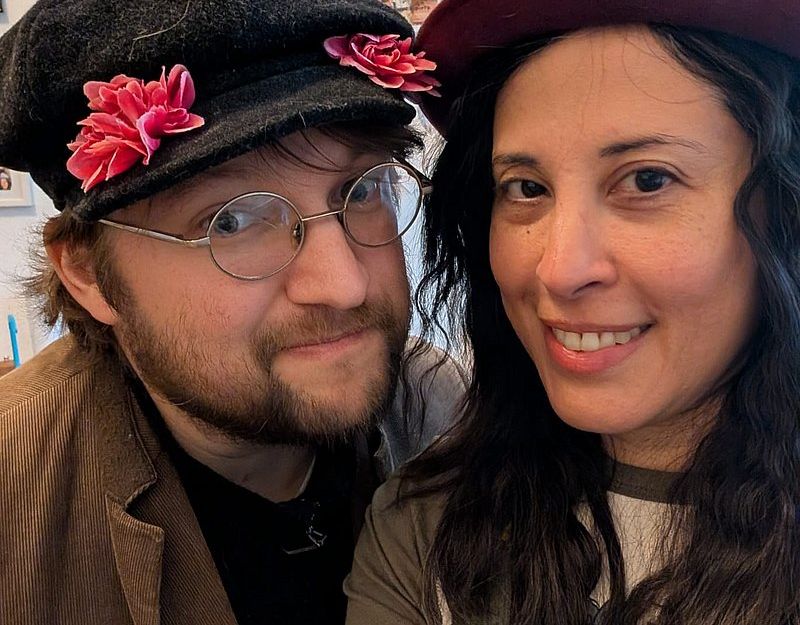 Zwei Personen mit Hüten, einer mit einem Apfel auf dem Hut, lächeln in die Kamera.