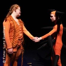 Zwei Personen in orangefarbenen Kostümen stehen auf einer Bühne und halten sich die Hände. Eine Person trägt ein Kostüm mit schwarzen Spitzen.