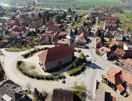 Luftaufnahme eines Dorfes mit einer Kirche im Zentrum, umgeben von roten Ziegeldächern und grünen Feldern im Hintergrund.
