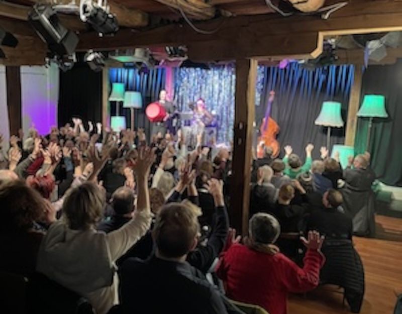 Ein Konzert in einem gemütlichen Raum mit Holzbalken, wo das Publikum die Hände hebt. Auf der Bühne stehen Musiker mit Instrumenten.