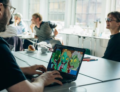 Person arbeitet an einem Laptop mit einer Zeichnung von Cartoon-Zombies, während andere im Hintergrund an einem Tisch sitzen und diskutieren.