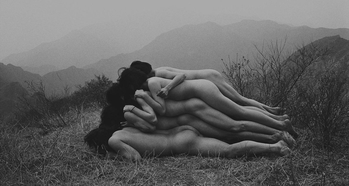 Eine Gruppe nackter Menschen liegt übereinander auf trockenem Gras in einer Berglandschaft.