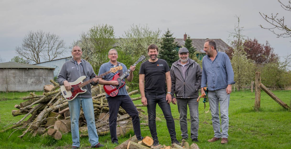 Fünf Männer stehen auf einer Wiese vor einem Stapel Holz, zwei halten Gitarren.