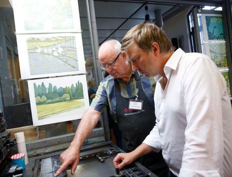 Zwei Männer arbeiten an einer Druckmaschine in einer Werkstatt mit Kunstwerken an der Wand.