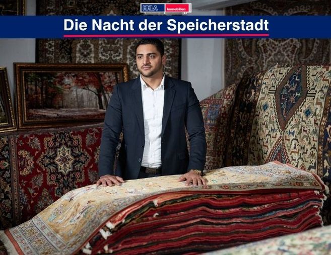 Ein Mann steht in einem Raum voller orientalischer Teppiche, die aufgestapelt sind. Im Hintergrund sind gerahmte Bilder zu sehen.