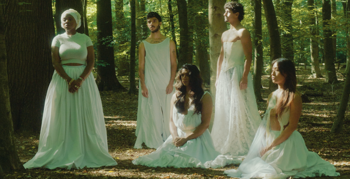 Fünf Performer*inne in weißen langen Kleidern posieren im Wald. Drei von ihnen stehen, zwei sitzen auf dem Boden. Keine*r schaut in die Kamera.