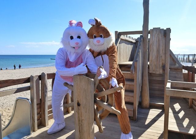 Zwei Personen in Osterhasenkostümen stehen auf einem Holzspielplatz am Strand.