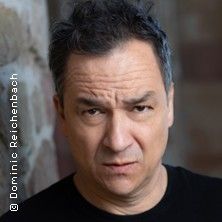 Porträt eines Mannes mit ernstem Gesichtsausdruck vor unscharfem Hintergrund.