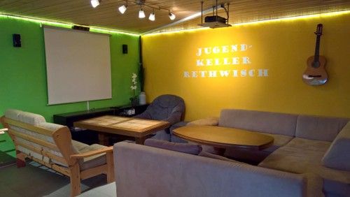 Ein gemütlicher Raum mit Sofas, einem Tisch, einer Leinwand und einer Gitarre an der Wand. Der Raum ist in Grün und Gelb gestrichen.