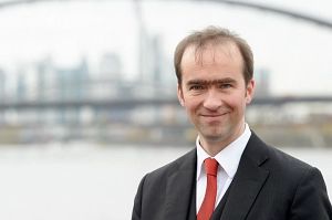 Ein Mann in Anzug und roter Krawatte steht vor einer verschwommenen Stadtlandschaft mit Brücke.