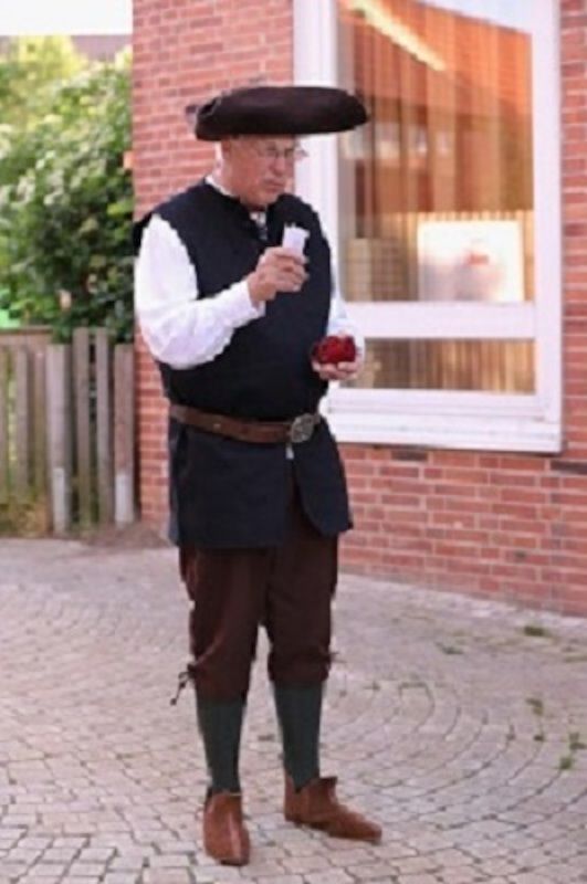 Ein Mann in historischer Kleidung steht auf einem gepflasterten Weg vor einem Backsteingebäude.
