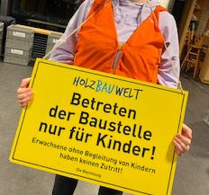 Eine Person hält ein gelbes Schild mit der Aufschrift: 'Betreten der Baustelle nur für Kinder! Erwachsene ohne Begleitung von Kindern haben keinen Zutritt!'