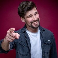 Ein Mann mit Bart und dunklem Haar zeigt lächelnd mit dem Finger auf die Kamera. Er trägt ein dunkles Hemd über einem weißen T-Shirt vor rotem Hintergrund.