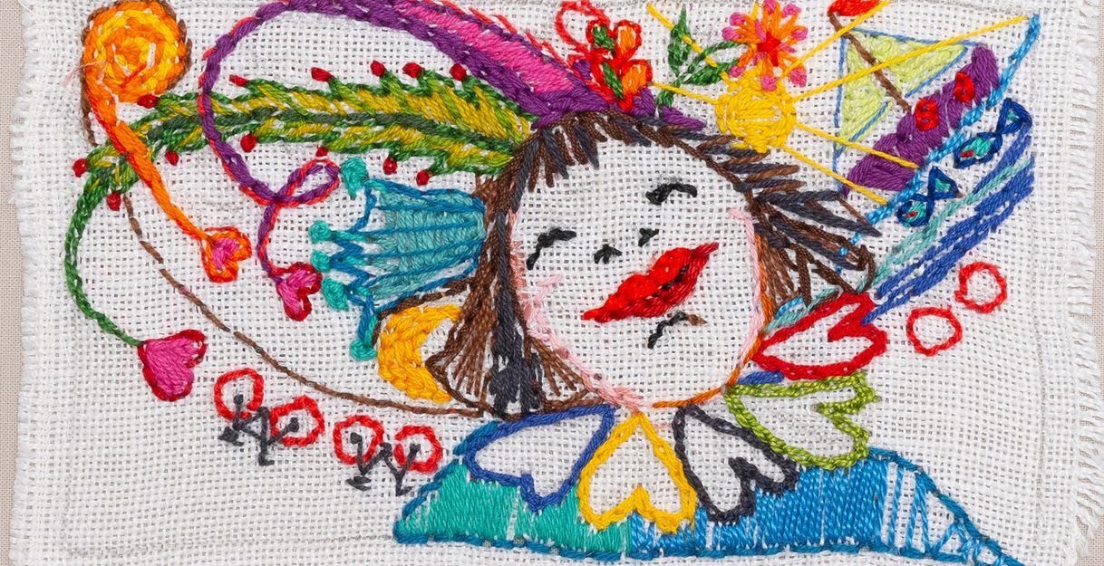Bunte Stickerei auf weißem Stoff mit einem stilisierten Gesicht und floralen Mustern.