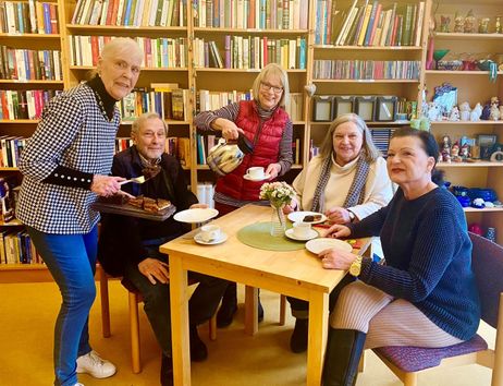 Eine Gruppe älterer Menschen sitzt an einem Tisch in einem Café mit Bücherregalen im Hintergrund. Eine Frau serviert Kuchen, eine andere schenkt Kaffee ein.