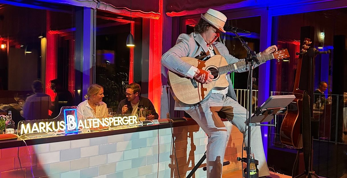 Ein Musiker spielt Gitarre auf einer kleinen Bühne in einem stimmungsvoll beleuchteten Raum.