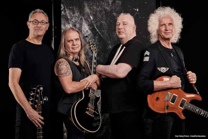 Vier Männer mit Gitarren und Schlagzeug posieren vor einem dunklen Hintergrund.