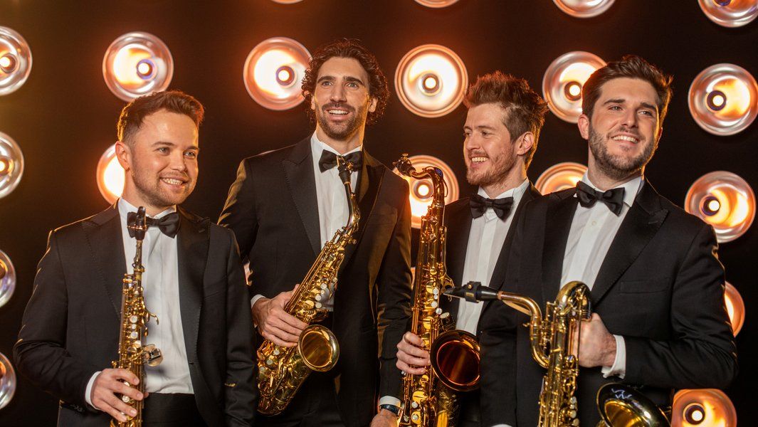 Vier Männer in Anzügen mit Saxophonen vor einer Wand aus runden Lichtern.