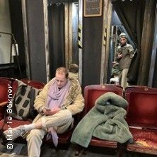 Zwei Personen in einem Raum mit Spiegeln und roten Stühlen. Eine Person sitzt, die andere steht im Hintergrund und schaut auf ein Handy.