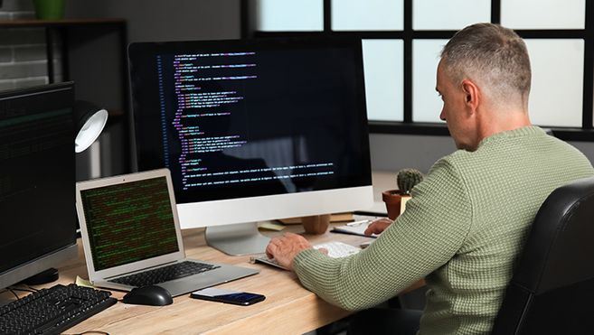 Es ist ein Mitarbeiter vor einem großen Monitor sowie einem Laptop zu sehen