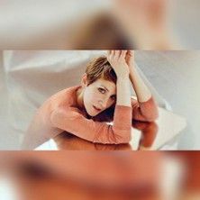 Eine Person mit kurzem Haar in einem orangefarbenen Oberteil posiert mit den Armen über dem Kopf.