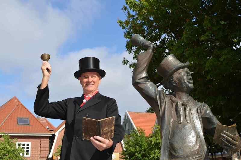Ein Mann in historischer Kleidung mit Zylinder und Glocke steht neben einer Statue in ähnlicher Pose in Otterndorf.