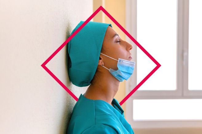 Eine Frau mit dem Kopf angelehnt an eine Wand mit einer grünen Haube und einem Mundschutz vor dem Kinn hat die Augen geschlossen.