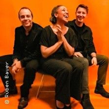 Drei Personen sitzen vor einem orangefarbenen Hintergrund und lachen.