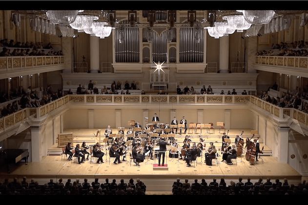 Orchester spielt in einem großen Konzertsaal mit Publikum auf Balkonen.