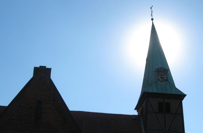 Kirchturm mit Uhr vor strahlend blauem Himmel und Sonne im Hintergrund.