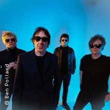 Vier Männer in dunkler Kleidung und Sonnenbrillen stehen vor einem blauen Hintergrund.